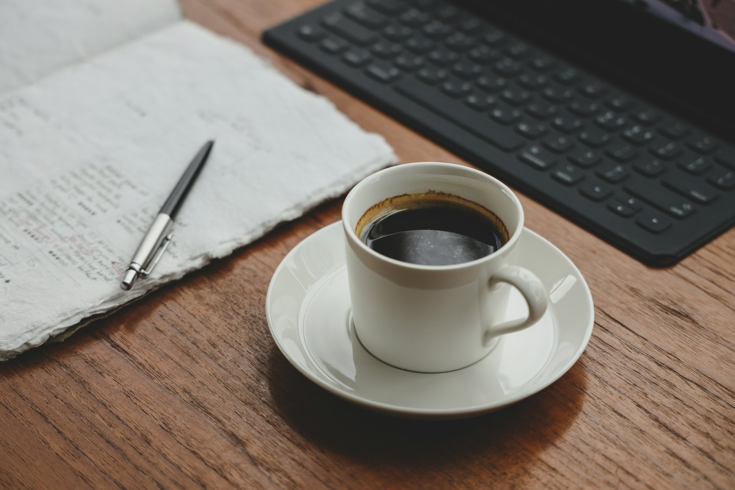 A white coffee cup sits on a desk next to an open notebook and pen.